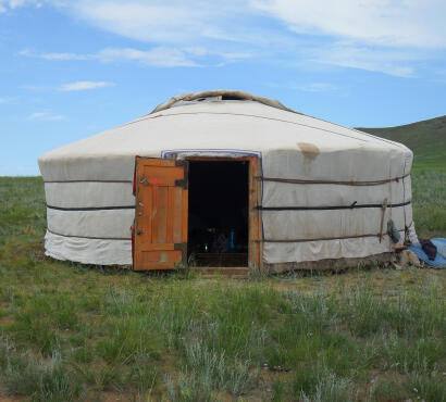 A
fully assembled traditional Mongolian ger covered in white canvas with an orange wooden door open, on a green grassy steppe.