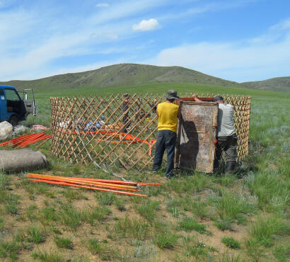 Three men arranging wooden lattice wall sections into a circle and fitting them to a door frame to assemble a Mongolian ger.
