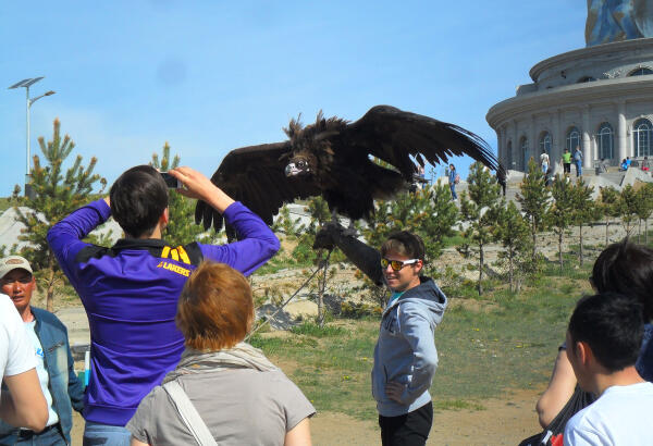 A tourist holds a large eagle with spread wings, with the base of equestrian statue visible in the background.